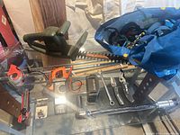 Overview of entire lot on glass table: hedge trimmer, clamp meter, tape, battery case, ratchet, wrench, pliers, multi-tool, utility knife, blue bag