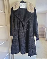 Full view of black textured long coat with fur collar on hanger