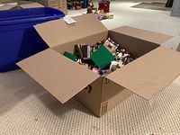 Cardboard box filled with loose LEGO pieces beside blue storage bin