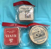 Three sealed plastic bags each containing a stack of vintage German advertising drink coasters: top square beige with German text, bottom left square red Manns, bottom right round München 1972 Olympic Münchner Bier