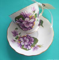 Teacup resting on saucer, showing violet floral decoration, gold rim, and candle wax inside cup