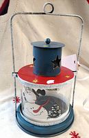 Front view of tealight lantern showing handle, red lid, blue chimney with star cutouts, glass cylinder with snowmen and trees