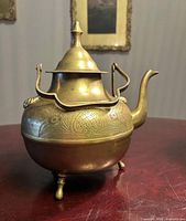 Front view of brass teapot showing spout, swing handle, etched decoration, tripod feet