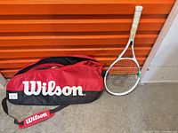 Wilson red and black tennis bag and white Jadnus racket leaning against wall