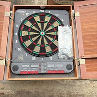 Cabinet open showing dartboard, scoreboard, plastic-wrapped darts