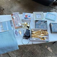 Overall lot on table showing curtain, utensil sets, cup warmer box, decorative figure, alarm clock, flatware, Bombay boxes