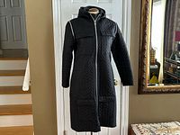 Front view of black quilted long coat on mannequin, hood visible, full length
