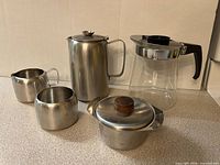 Group shot of five kitchen items: stainless coffee pot, creamer, sugar bowl, lidded dish, glass carafe