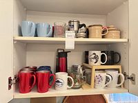 Cabinet shelves showing assorted ceramic mugs, clear glass mugs, insulated travel mug, beige sugar pot, shelf riser, glass teapot