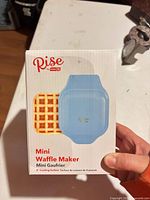Front of box showing Rise by Dash light blue mini waffle maker