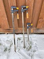 Three bar clamps leaning against wooden fence in snow