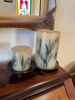 Two pillar candles with embedded botanical designs on individual metal pedestal stands