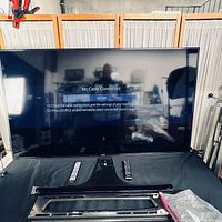 Front view of powered-on 65" Samsung TV with two remotes and wall-mount bracket visible