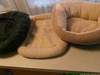 Three dog beds on table: dark grey round, cream rectangular pad, beige oval bolster