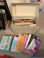 Lot overview showing storage box filled with greeting cards, separate stack of cards and boxed set