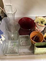 Plastic bin with clear glass vases, red hobnail vase, terracotta pot, square glass vase, ribbed glass vase, green embossed metal planter, striped ceramic pot with faux flowers