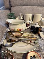 Pile of flatware on green-rim plate, fruit motif gravy boat, small cream tumblers, green glass cups, dachshund figurine visible