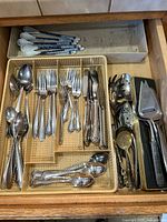 Drawer view showing assorted silver-tone flatware in tray, ceramic-handled spoons, boxed serving utensils including pie server, slotted spoon, spaghetti server, cheese plane