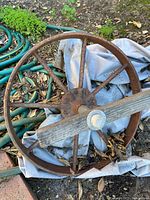 Side view of rusted metal spoked wheel with wooden strut attached, garden hose visible but excluded