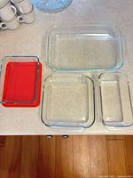Top-down view of five glass cookware pieces including large and medium baking dishes, loaf pan, and two small storage containers with red lids on countertop.