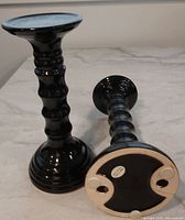 Two glossy black ceramic pillar candle holders, one standing upright the other lying flat showing underside pads