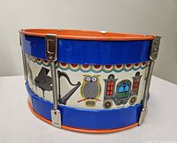Side view showing blue tin drum with owl, piano and harp illustrations