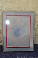 Framed embossed painting "Blue Plate Special" by John Kalamaras leaning against a textured wall, showing front view with glare reflections on glass.