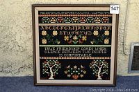 Framed needlepoint wall art with alphabet, numbers, floral and tree motifs, and a friendship quote on dark fabric background.