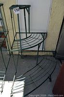 Green metal outdoor plant stand with two curved tiered shelves showing front and side view, placed on concrete outside a building.