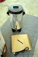 Photo of stainless steel WestBend coffeemaker with black lid, side handles, front spout on a grey carpet along with power cord.