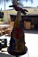 Full view of the conical African ceremonial hat with colorful beadwork and bird atop on wooden pedestal stand, outdoors.