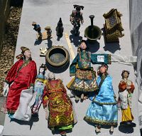Bird’s-eye view of seven dolls, brass bowl, mortar and pestle, brass shrine, brass elephant and carved figurines on white cloth.