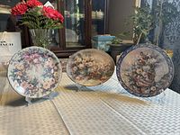 All three Bradford Exchange plates on stands: round rose wreath, oval bouquet, round blue-border pansy basket
