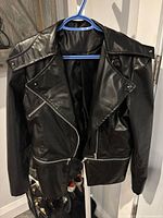Front view of black faux leather moto jacket on hanger showing asymmetrical zipper and detachable panel