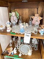 Two-shelf cabinet view showing variety of ceramic, glass and fabric angel figures, doily, and bear