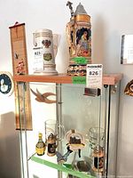Overall lot on display cabinet showing two large steins on top, two glass mugs and miniatures inside