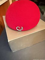 Red felt hat resting on box, view of exterior and decorative pin