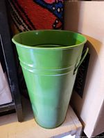 Tall bright green painted metal bucket with single side handle loop, shown on shelf between other items