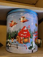 Front view of round metal Christmas tin showing gingerbread house, candy canes, elves, trees, presents, blue lid