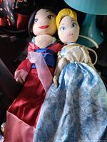 Two princess plush dolls side by side in vehicle trunk; shows overall appearance, clothing colors, facial embroidery, size relation