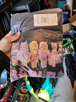 Sealed package held in hand displaying black T-shirt with multicolor character graphic and size label