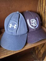 Gray and brown Under Armour baseball caps side by side showing front logos and brim condition