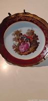 Front view of Limoges plate showing burgundy rim, gold scrolls, courting couple scene, stand attached