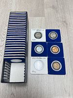 Stack of 21 blue coin holders inside black storage box alongside five of the six coins in holders