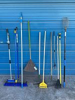 Full view of 11 long-handled garden and cleaning tools against blue wall