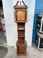 Full front view of wooden floor clock showing dial, shelves, and base cabinet