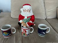Santa cookie jar with three holiday mugs on sofa