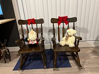 Two wooden children’s rocking chairs side by side with plush Santa and teddy sitting on seats