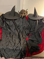 Two black witch costumes with hats and brown monster mask displayed on red surface