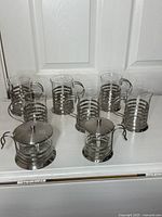 Eight-piece glass and stainless-steel coffee set arranged on surface, showing cups in back row and sugar and creamer in front